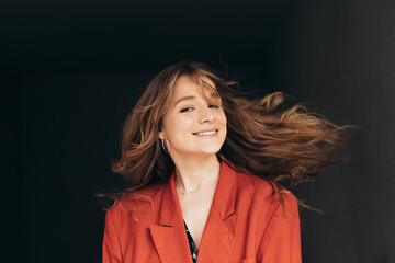 Young beautiful amazing woman in orange jacket waves her hair and poses on camera. Girl stand alone in studio. Isolated over dark background. Nice attractive model on picture.