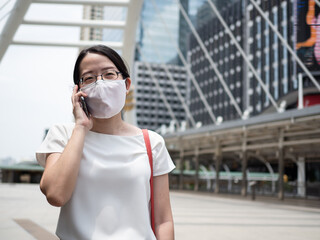 Beautiful Asian women wearing disposable medical face mask, using smartphone while in a public area, roadside, or city center, as new normal trend and self-protection against Covid19 infection.