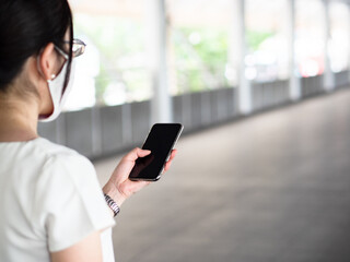Beautiful Asian women wearing disposable medical face mask, using smartphone while in a public area, roadside, or city center, as new normal trend and self-protection against Covid19 infection.
