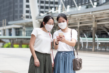 Two Asian women wearing medical face mask every time outside the house, as new normal trend and self-protection against Covid19 infection, using smartphone for selfie or finding business information
