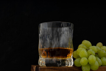 A glass of whiskey and a bunch of white grapes on wooden table against dark background in pub