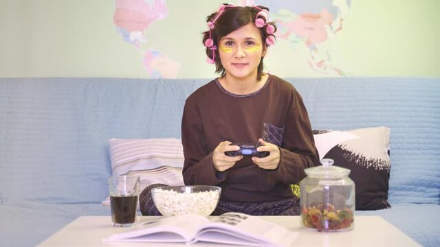girl plays console, holds joystick in hands, eats popcorn, sits on sofa on table marmalade and glass of soda. young woman wears brown pajamas, patches under eyes, hair curlers. High quality 4k footage