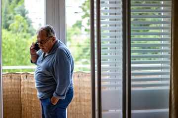 Worried senior grandfather talking with phone near the window at home. Senior man talking using mobile phone, appears to be unhappy, sad or angry.