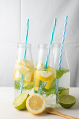 Top view of three bottles with blue straw, water and lemon, lime and mint slices, on white table with half lemons and wooden spoon, white background, vertical