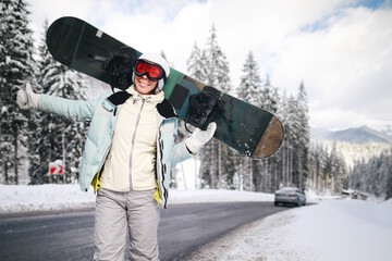 Young woman snowboarder hitchhiking on a winter road with beautiful snowy mountains in background