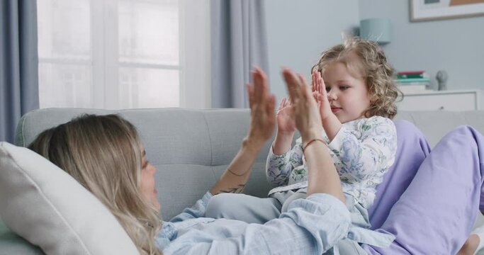 Middle plan of happy parent and cute kid playing patty cake at home, carefree young mother and little doughter lap hands having fun together laughing enjoying funny game relaxing on couch.