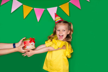 Young birthday girl in yellow blouse with cap taking a present from her mom's hand on green background.