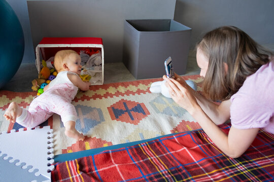 Young mom with smartphone taking picture of her curious baby daughter checking box with stuff, searching for her toys. Childhood or child care at home concept