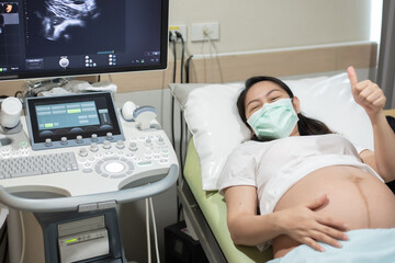 .Smiling Asian pregnant woman with white T-shirt lies at the hospital bed and rise her hand thumbs up before ultrasound