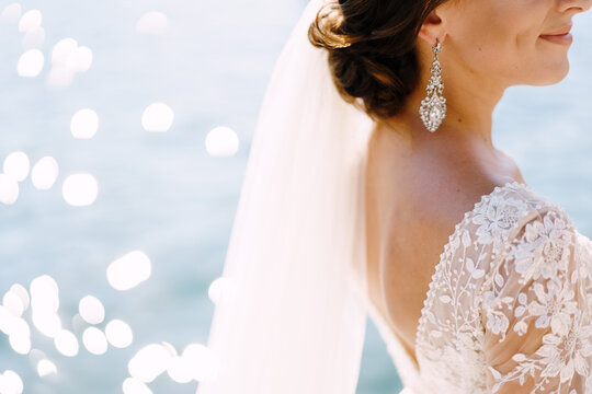 Close-up of the neck and neck of the bride, face profile. Beautiful big earring in ears, shoulder with white lace dress on background of glare of water. Fine-art wedding photo in Montenegro, Perast.