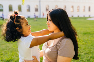 Obraz premium Cutie curly mexican dark skinned preschooler girl playing, hugging her latin mother outdoor in european city park. Growing up, leadership, copying female psychology behavior, tenderness concept.