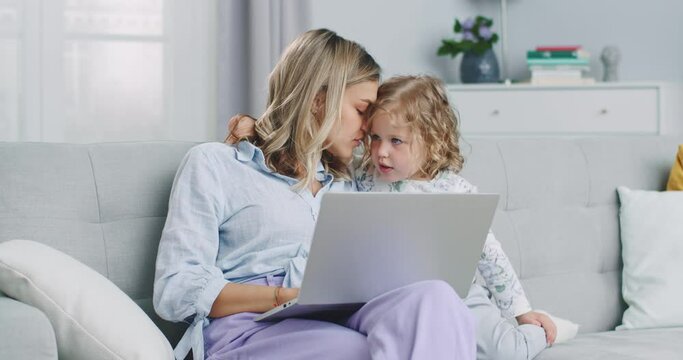 Middle plan of mother multitasking using laptop computer at home with her baby. Candide genuine and real life mom works and parenting. Self-isolation. Concept of family, parenthood, togetherness.