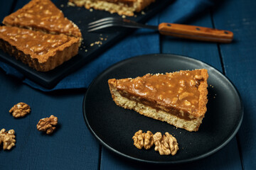 A piece of tart on the black plate and chopped walnut caramel pie on the wooden table