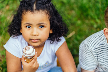 Top view portrait of cute hispanic dark skinned curly sweet girl eating ice cream, sitting on the green grass at park outdoor. Childhood, summer vacation and healthy food concept.