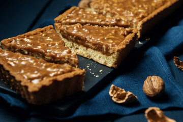 Chopped walnut-caramel tart on a black serving board with walnuts on wooden table
