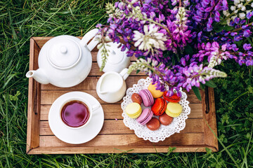 A cozy breakfast in the fresh air. A vase with lupine flowers, a teapot and a cup of coffee tea, a cake with macaroons on a wooden tray. Mother's Day or Women's Day. 