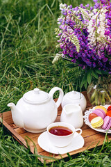 A cozy breakfast in the fresh air. A vase with lupine flowers, a teapot and a cup of coffee tea, a cake with macaroons on a wooden tray. Mother's Day or Women's Day. Soft selective focus.