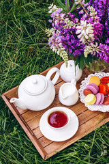 A cozy breakfast in the fresh air. A vase with lupine flowers, a teapot and a cup of coffee tea, a cake with macaroons on a wooden tray. Mother's Day or Women's Day. Soft selective focus.