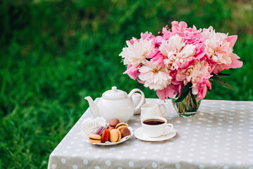 A cozy romantic breakfast in the open air. Peony flower vase, teapot and cup of tea, macaroon cake on the table. Congratulations on mother's day or women's day. 