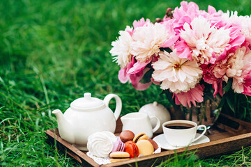 Cozy breakfast outdoors. Peony flower vase, teapot and cup of tea, macaroon cake on a wooden tray. Congratulations on mother's day or women's day. 