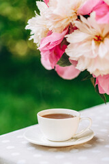 A cozy romantic breakfast in the open air. A vase of peonies flowers, a cup of coffee tea on the table. Soft selective focus.