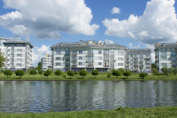 Fototapeta na wymiar Residential blue and white elite apartment buildings on the river bank near the park with a trimmed lawn and trees with round crowns. Summer River embankment, huge white clouds.