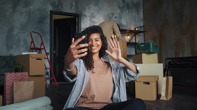 Multiracial friends moving into new house. Guy unpacking indoor plants. Girl smiling, waving hand and recording video for her blog using smartphone