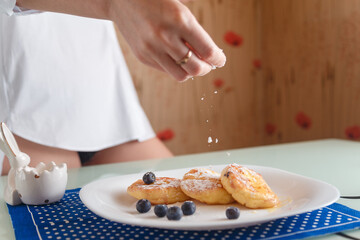 Obraz na płótnie Canvas Female hand sprinkling powdered sugar on traditional syrniki or cottage cheese pankaces with blueberries and honey on white plate with blue napkin on kitchen table. Healthy breakfast, diet concept.