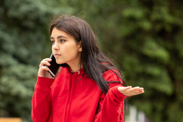 Dark-skinned indignant woman in red spreads her arms while talking on the phone, on the street