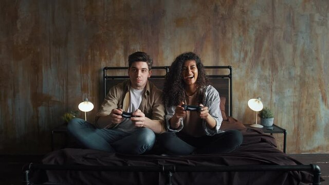 Excited man and woman in casual outfit are laughing, holding wireless gamepads and playing video game on console. Sitting on bed in bedroom