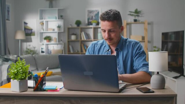 Cheerful young man is typing using laptop in the living room at his desk. And snaps his fingers.
