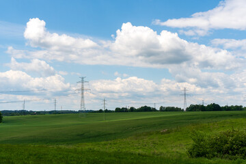 High voltage lines and power pylons in a flat and green agricultural landscape on a sunny day with cirrus clouds in the blue sky.