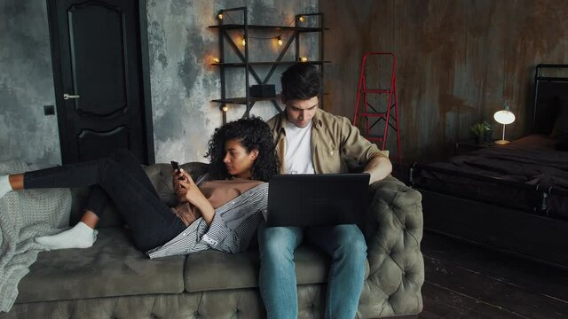 Guy and girl moving to a new house. He working at laptop computer, she using mobile phone. They sitting on gray couch in room with cardboard boxes