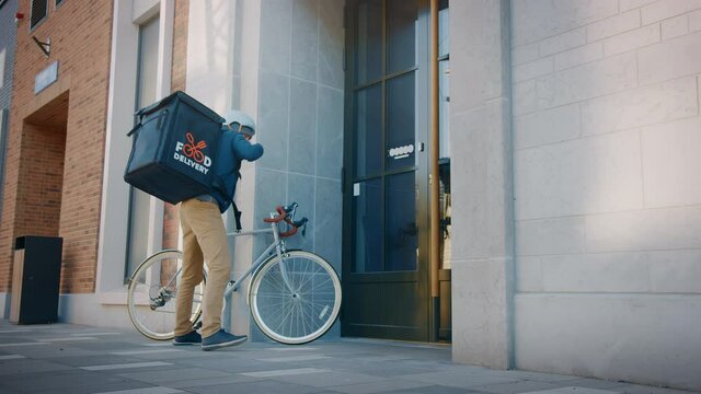 Food Delivery Man Wearing Thermal Backpack Rides Bike and Delivers Restaurant Order, Leaving it Under the Door. Concept: Contactless (No Contact) Delivery for Social Distancing, Self Isolation