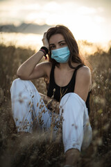 Young woman with black hair wearing a mask for health care in a field at the evening surrounded by...