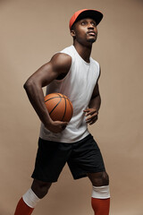 photo of a dark-skinned athletic basketball player in studio on a beige background posing with a ball, wearing a white t-shirt, red cap, black shorts and he is looking up