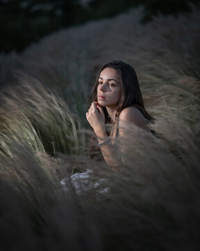 Young woman with long black hair standing between plants looking at the camera during night time after the sunsets with a light hitting her face