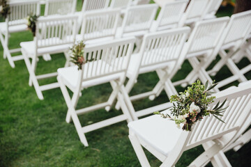White wooden chairs stand on the green grass in the wedding ceremony area, on the chairs there are decorative compositions of flowers and greenery