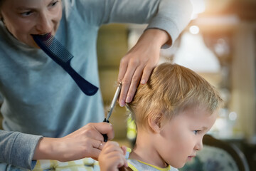 Obraz na płótnie Canvas Close-up young adult caucasian mother making haircut fot cute adorable son toddler boy at home due quarantine and lockdown. Mom cutting hair of child with scissors and hairbrush