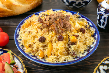 National Uzbek pilaf with meat, achichuk salad of tomato, cucumber, onion in plate with traditional pattern, cilantro, cherry tomatoes, garlicbread tortilla - patir on dark wooden table Top view