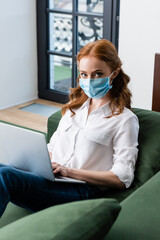 Selective focus of red haired woman in medical mask looking at camera while using laptop on couch