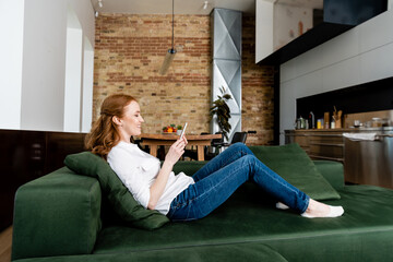 Side view of smiling woman using smartphone on couch at home