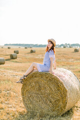 Naklejka na ściany i meble Young brunette Caucasian girl wearing straw hat and blue striped dress, sitting on a hay bale and posing at camera. Summer portrait of young woman in beautiful wheat harvesting field at sunset