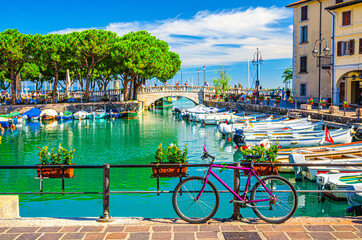 Bicycle bike near fence of old harbour Porto Vecchio with motor boats on turquoise water and Venetian bridge in historical centre of Desenzano del Garda town, blue sky, Lombardy, Northern Italy - obrazy, fototapety, plakaty