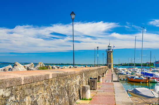 Stone pier mole with lighthouse, street lights and yachts on boat parking port marina in Desenzano del Garda town, blue sky white clouds background, Lombardy, Northern Italy