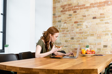 Selective focus of woman holding toast and using laptop near fruits on table