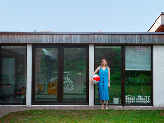 A pretty girl in a summer dress poses against the background of the pool window in the backyard with an inflatable ball.