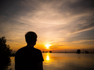 Fototapeta premium Silhouette of a man with a short haircut on the background of the lake in the light of the setting sun. Copyspace. Warm shades.