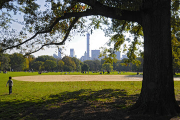 Vue sur central park avec des gens et des enfants jouant au ballon sur le gazon et à l'ombre des branchages en automne, sur fond de gratte-ciel à l'horizon.