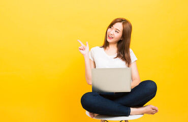 Asian happy portrait beautiful cute young woman teen smiling sitting crossed legs on chair with laptop computer point finger side looking to side isolated, studio shot yellow background with copy spac